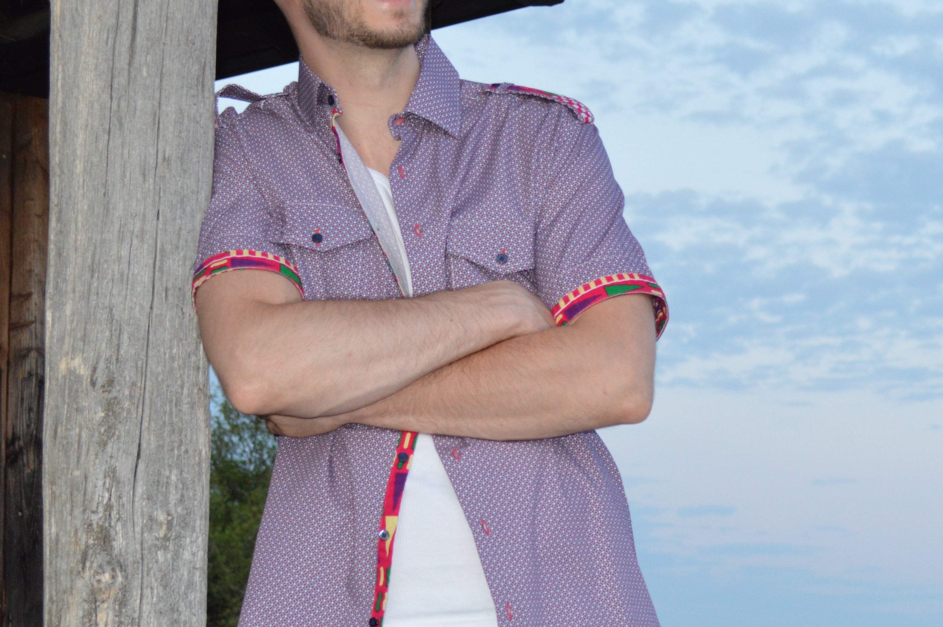 Model possiet angeleht an einer Holzhütte mit einem Kurzarm Hemd.