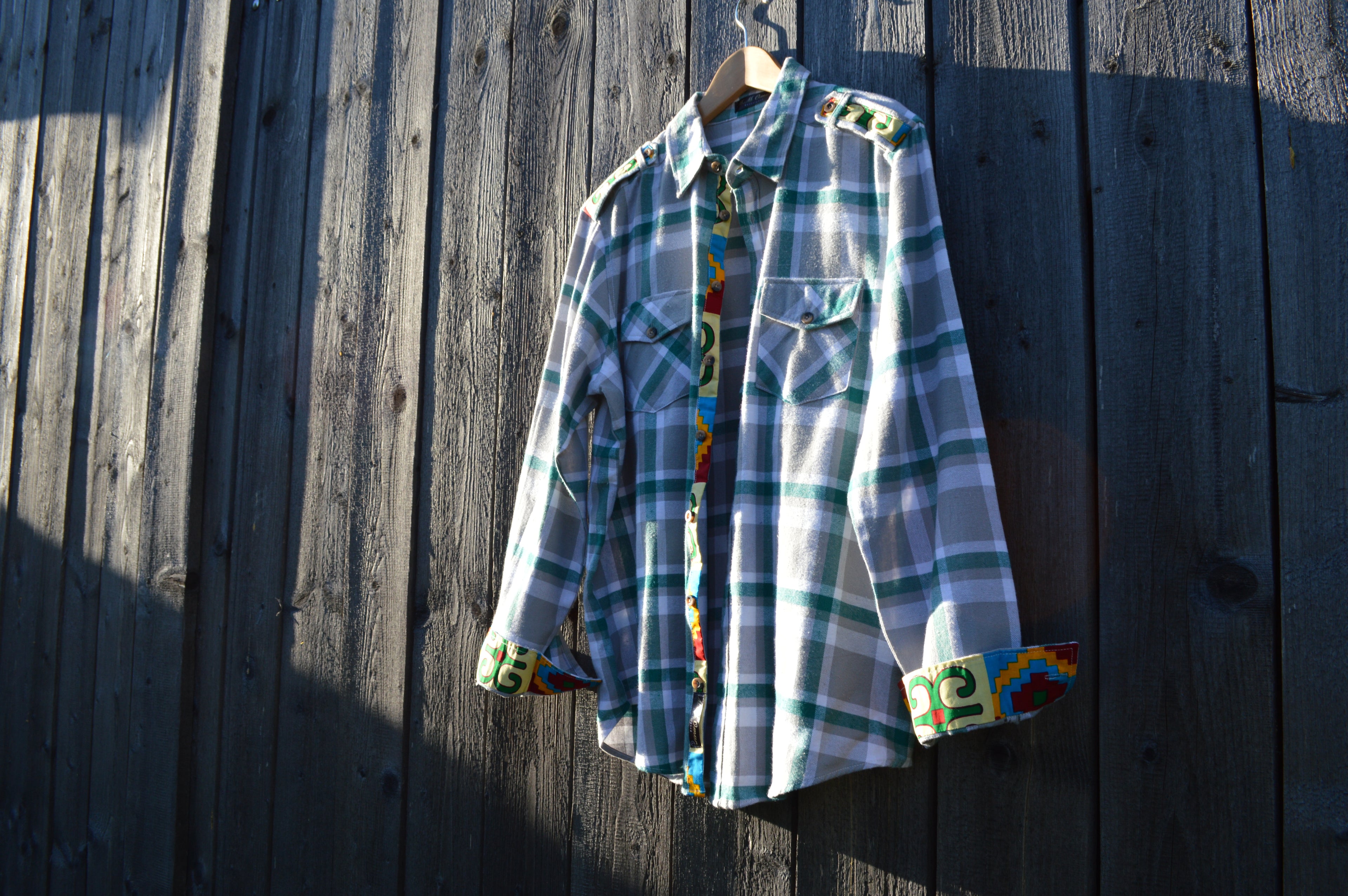 Karriertes Flanellhemd mit Afrikanischen Waxdruckmustern an Manschetteninnenseite,Knopfleiste und Schulterklappen. Hängend präsentiert an Holzwand.